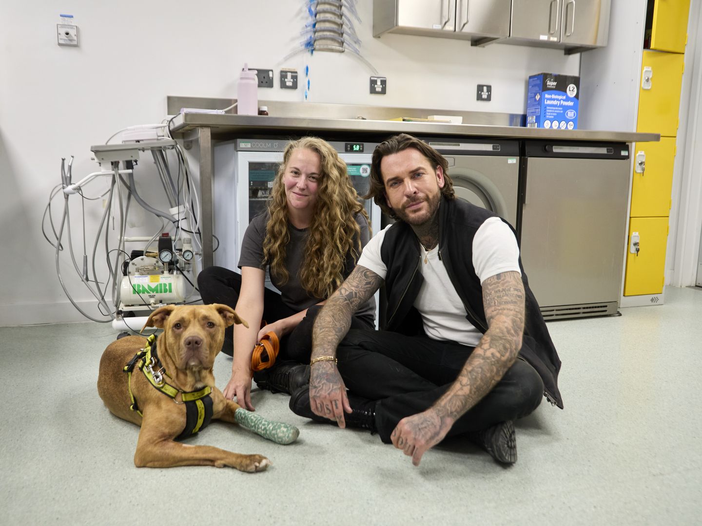 Pete wicks sitting down on the floor with a lady and a tan bully breed dog smiling at the camera